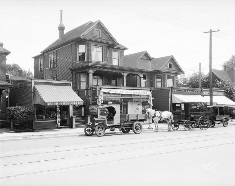 Delivery trucks and a horse-drawn wagon outside shops built onto the front of houses in the 1000 block of Denman Street