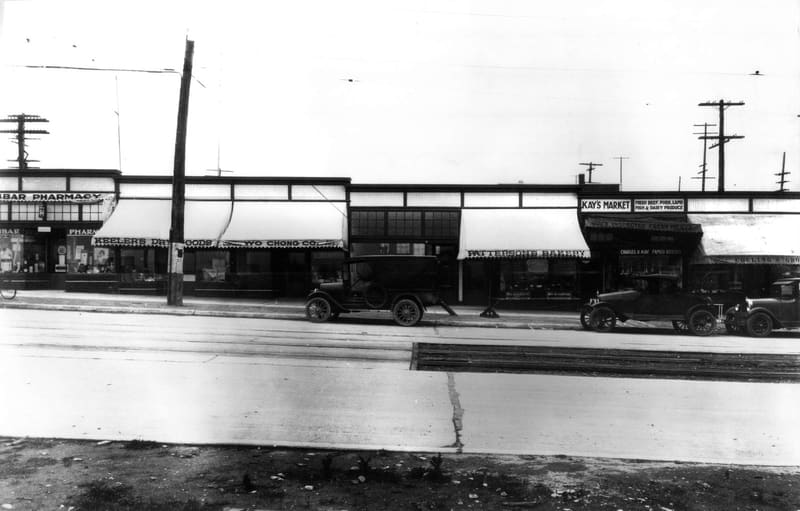 Shops on the west side of the 3200 block of Dunbar Street, among them Dunbar Pharmacy and Kay's Market, with cars out front